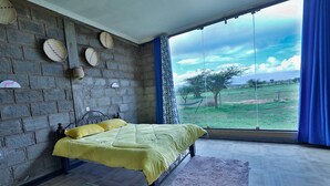 Ein Schlafzimmer mit großem Fenster, einem Bett mit gelber Bettdecke, einer Ziegelwand und Blick auf ein grünes Feld.