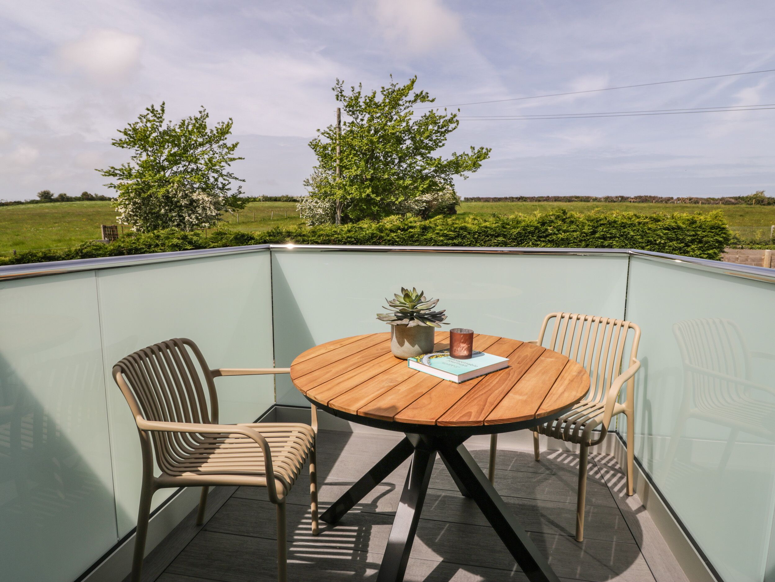 A corner balcony with a round wooden table, two chairs, a potted plant, and a book.