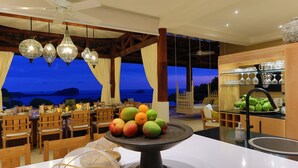 A dining area with a view of the ocean, wooden furniture, hanging lights, and a fruit display. - 6BR Luxury Ocean View Villa • Infinity Pool • Private Cinema • Spa (Quepos)