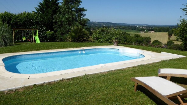 A rectangular swimming pool with a clear blue water, surrounded by a grassy area and trees. There are two lounge chairs and a swing set visible.