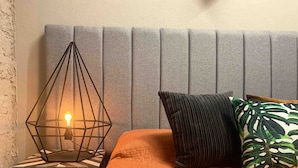 A bed with an orange quilt, a grey upholstered headboard, a black metal nightstand, and a geometric wire lamp.