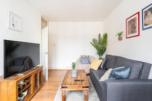 A living room with a flat-screen TV on a wooden stand, a grey sofa with patterned cushions, a coffee table, and a potted plant.