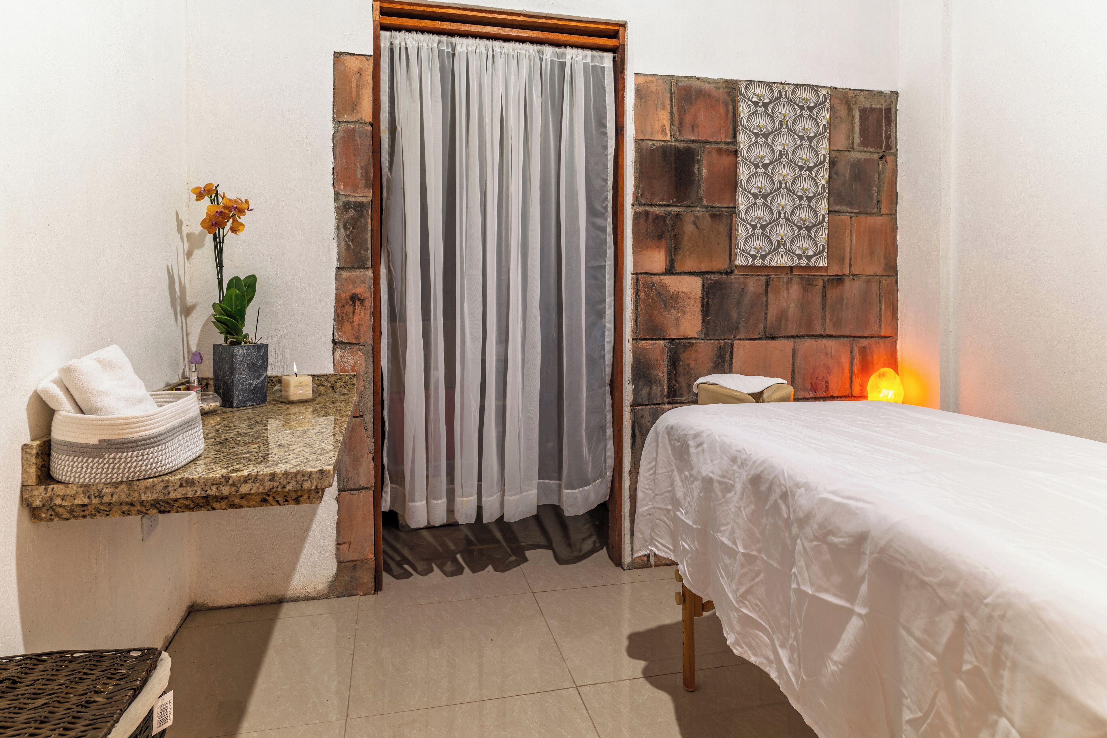 A room with a bed, a small sink, a towel, a plant, and a wall with a patterned tile.