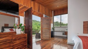 A spacious bedroom with wooden flooring, a large window offering a view of greenery, a wooden wardrobe, and a bedside table with a plant. - Hotel Ribera La Paz (Piedades Norte)
