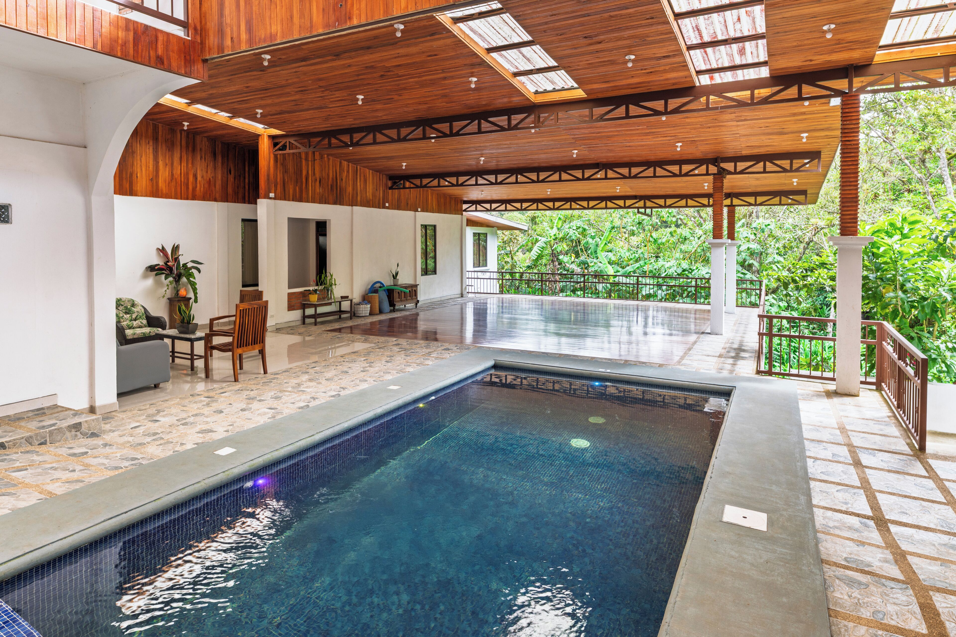 A swimming pool with a wooden deck, seating area, and a view of lush greenery.