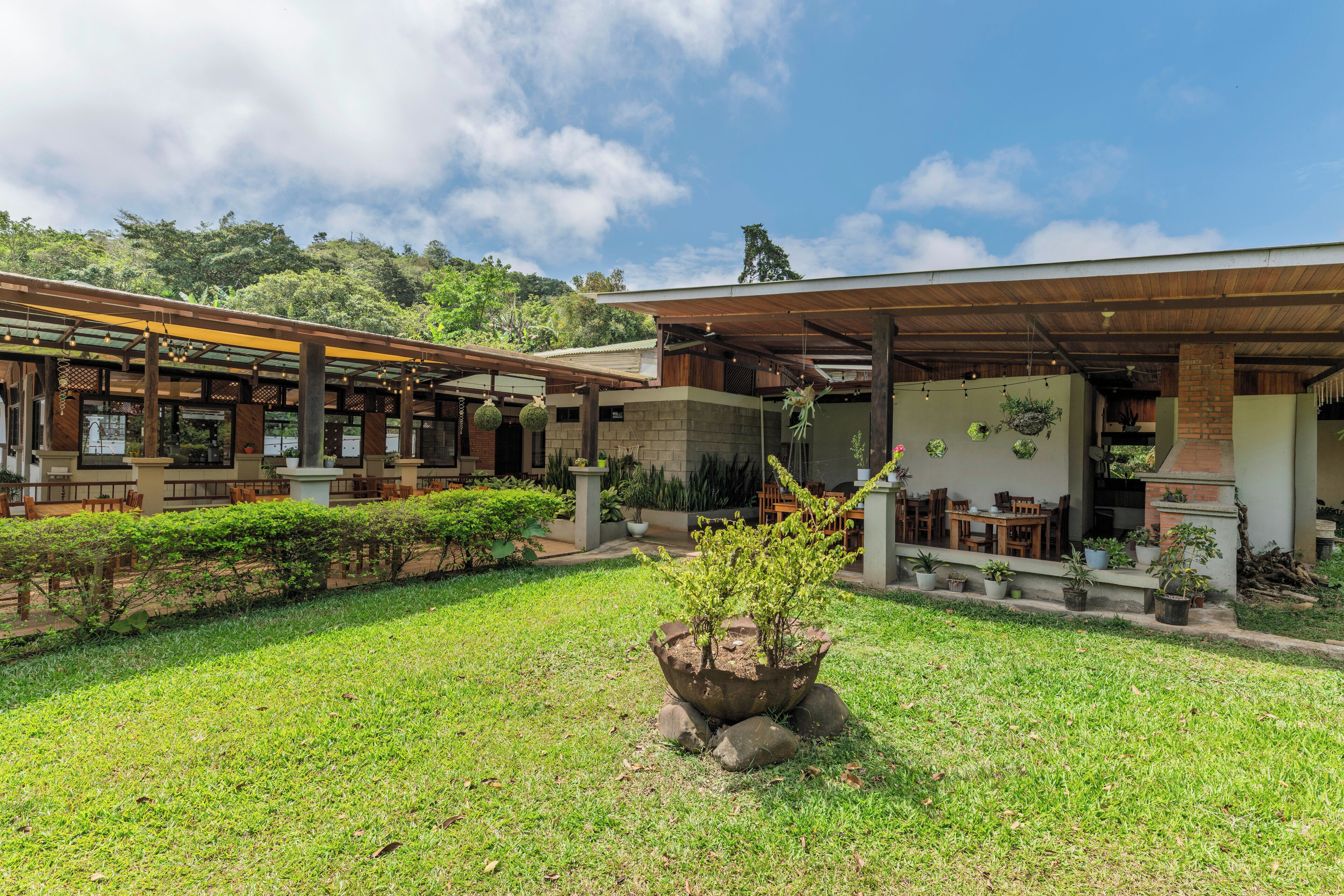A modern building with a covered outdoor seating area, a garden with a central planter, and a view of a lush hillside.