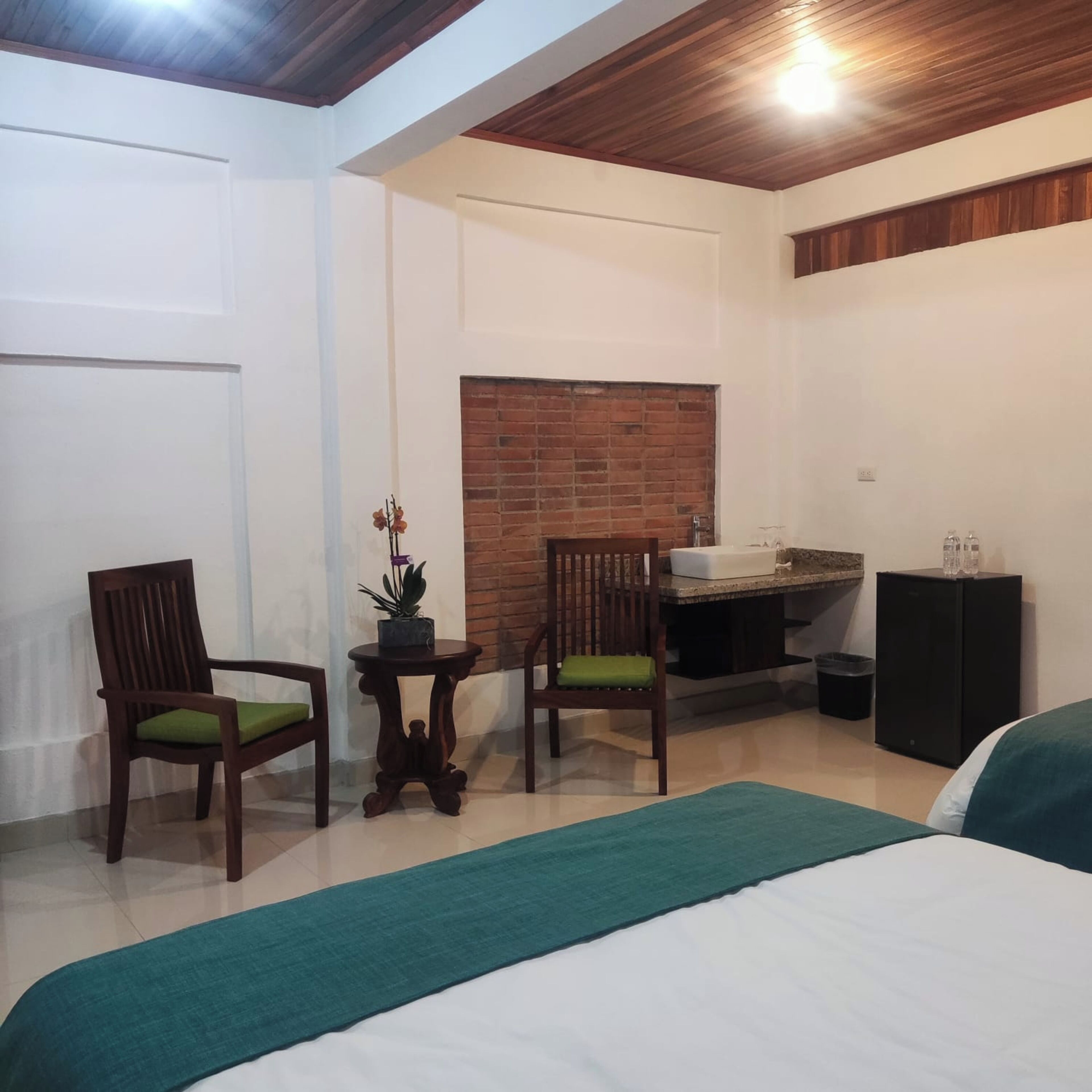 Une pièce comprenant un lit, deux chaises en bois avec des coussins verts, une petite table avec un pot de plante, un mur en briques et un coin lavabo.