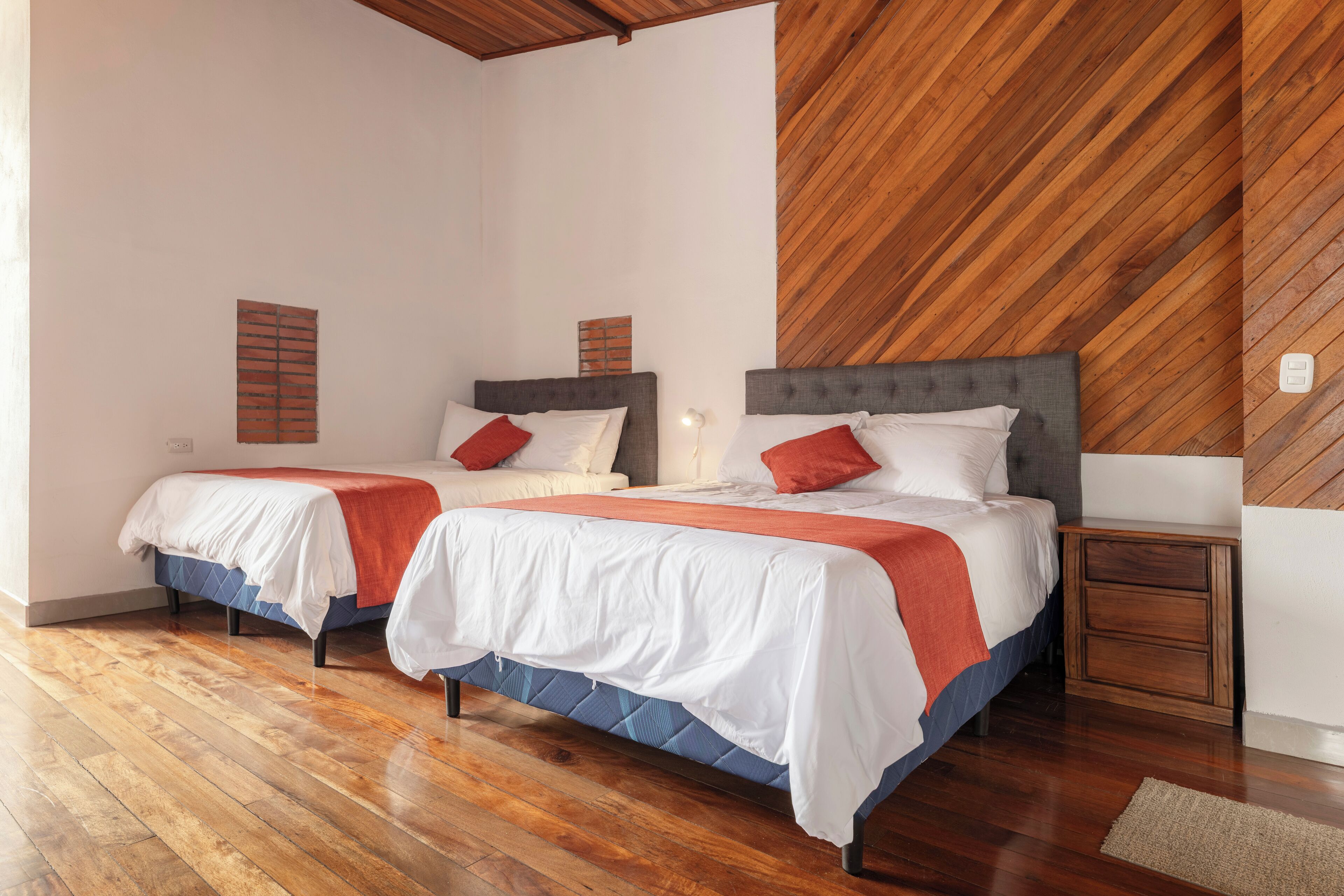 A bedroom with two beds, wooden flooring, a wooden accent wall, and a nightstand.