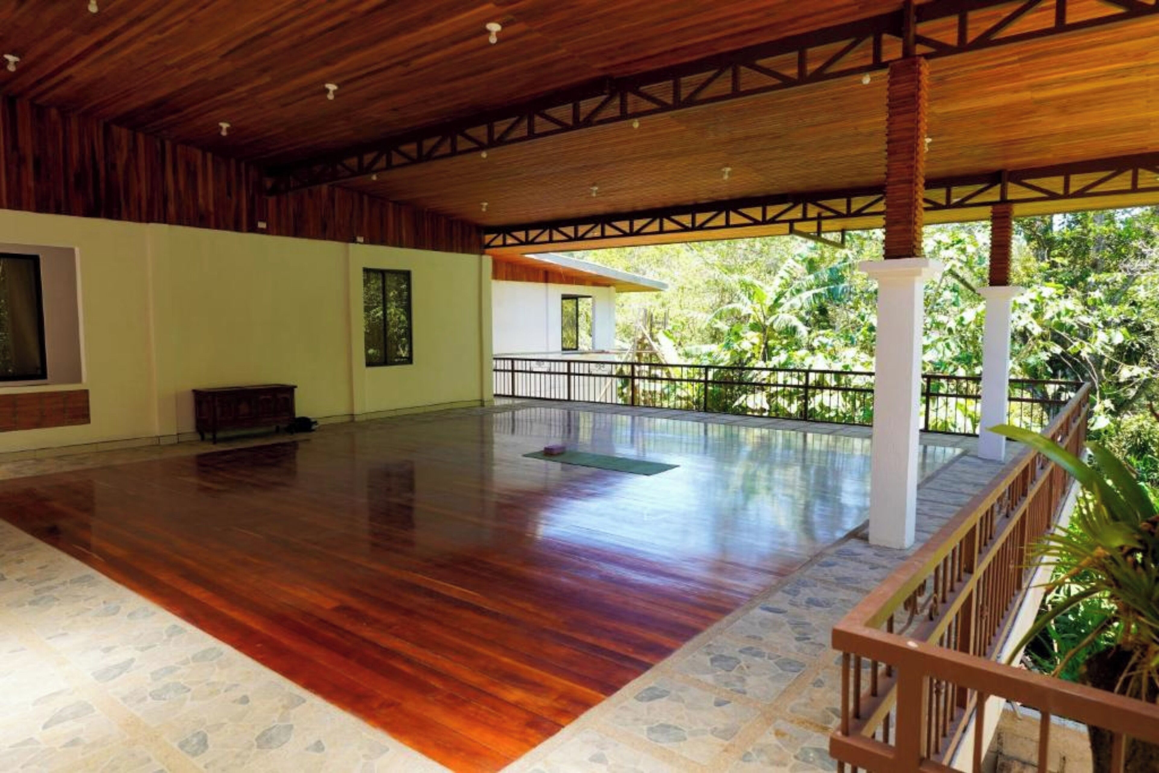 An indoor space with a wooden floor, a balcony with a railing, and a view of greenery outside.