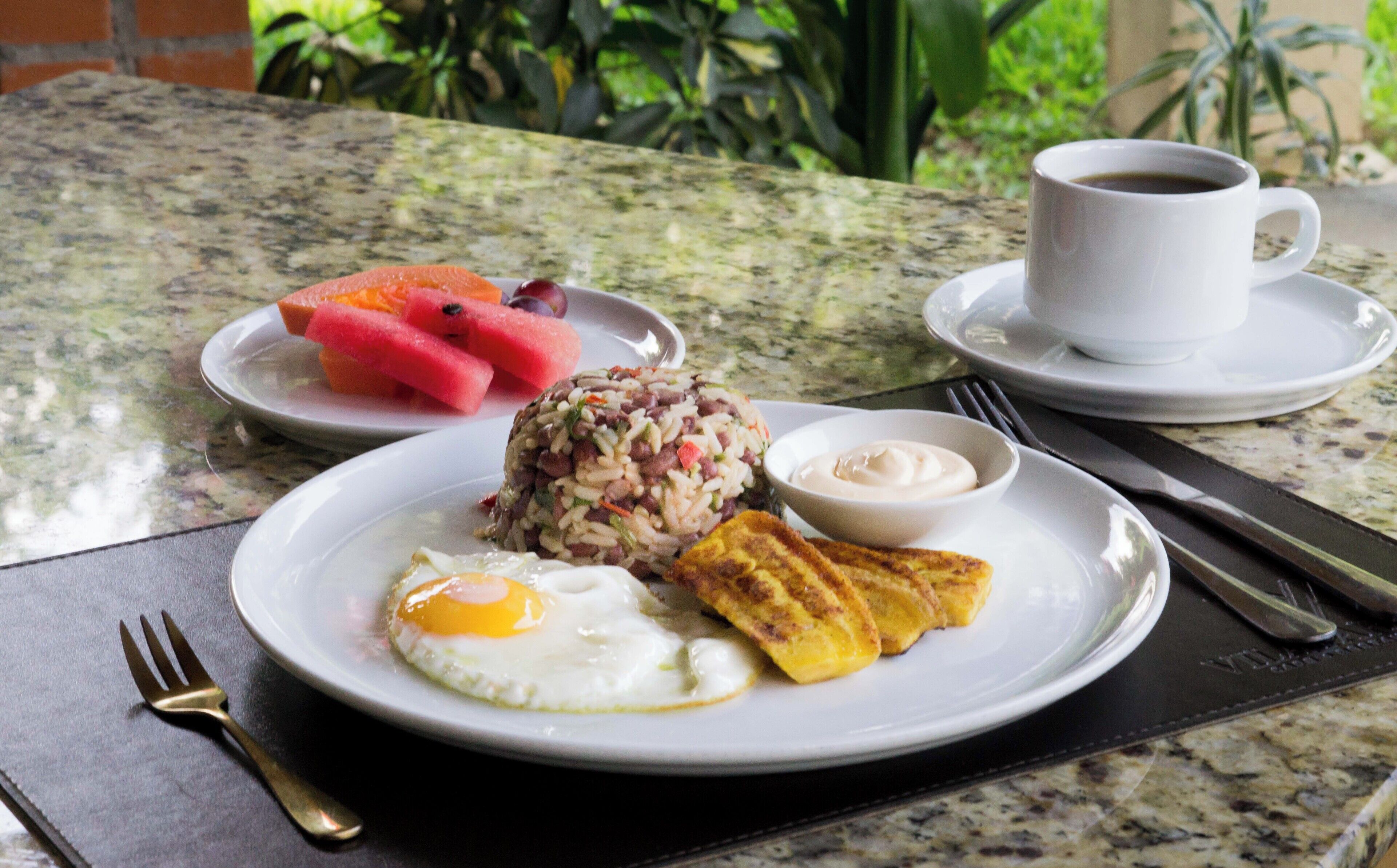 A breakfast plate with eggs, rice, beans, and plantains, a bowl of sauce, and a cup of coffee on a table.