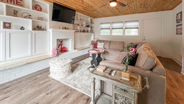 Un salon chaleureux avec une cheminée, une cheminée en briques blanches, un plafond en bois, une grande fenêtre et un canapé beige.