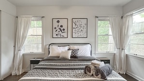 Une chambre à coucher avec un lit, deux fenêtres avec des rideaux, un ventilateur de plafond et deux tableaux accrochés au mur.