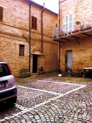 A cobblestone courtyard with a parked car, a brick building with a balcony, and a door.
