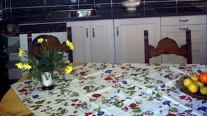Une cuisine avec des meubles blancs, un plan de travail noir et un lustre. On y trouve une table recouverte d’une nappe à motifs floraux et un vase rempli de fleurs.