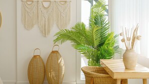 A room with a wooden dining table, two wicker vases, a macrame wall hanging, a potted plant, and a wicker chair.