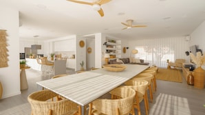 A spacious living area with a long dining table, wicker chairs, ceiling fans, and a kitchen in the background.