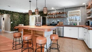 A modern kitchen with a central island, bar stools, hanging wooden light fixtures, and a green leafy wall. - 1 Block From W Main St - Quiet Spacious Getaway (Fredericksburg)