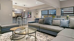 Un salon moderne avec un canapé gris, une table basse en verre et un coin cuisine à l’arrière-plan.