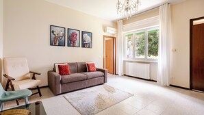 Un salon spacieux avec un canapé gris, un fauteuil beige et une table basse en verre. Trois tableaux encadrés ornent le mur, ainsi qu’un lustre et une grande fenêtre avec des rideaux.