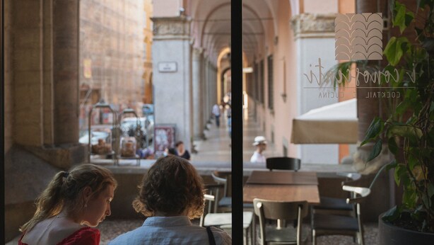 An outdoor café with tables and chairs, a stone building with arches, and a sign with a logo.