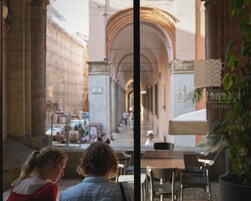 Un caffè all'aperto con tavoli e sedie, un edificio in pietra con archi e un cartello con un logo.