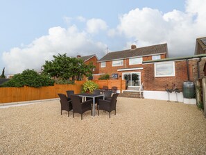 A patio with a table and chairs, a brick house, a wooden fence, and a gravel ground.