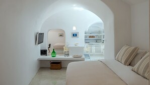 A minimalist room with a bed, a small table, a chair, a TV, and a kitchen area visible through an archway. - Crown Of Fira (Santorini)