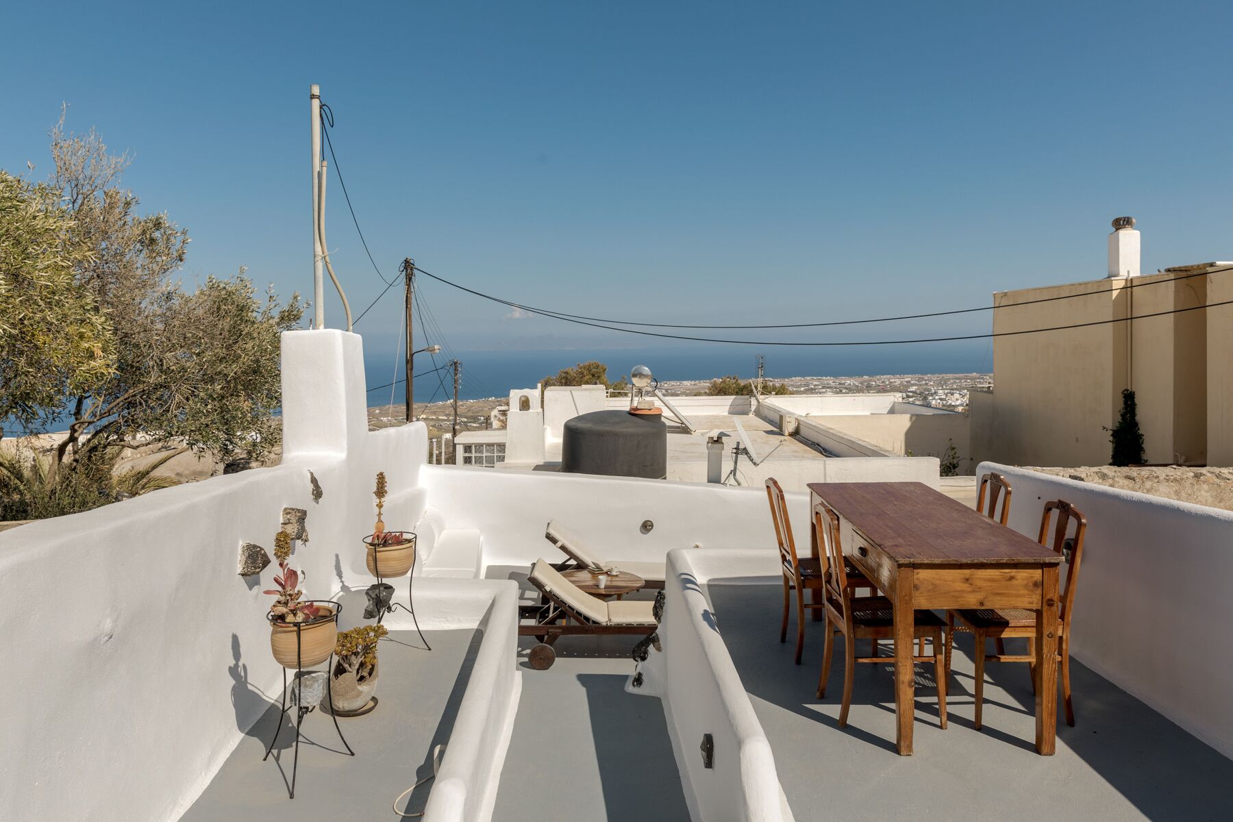 A rooftop terrace with a dining table and chairs, potted plants, and a view of the sea and cityscape.