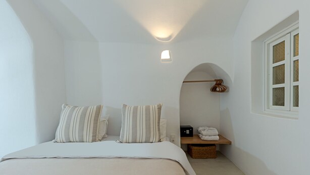 A bedroom with a bed, pillows, a bedside table, a lamp, a window, and a small seating area with a chair and a basket.