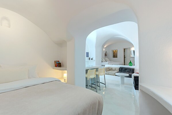 Quarto de hotel moderno com uma cama grande, uma pequena área de refeições com mesa e cadeiras, e vista para o quarto adjacente.