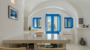 A dining area with a white table, blue doors, and a view of the outside. - Crown Of Fira (Santorini)