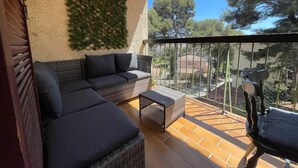 Ein Balkon mit einer Rattan-Couch, einem kleinen Tisch und einem Stuhl. Eine grüne Wanddekoration und der Blick auf Bäume und Gebäude sind zu sehen.