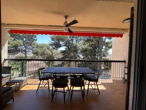 Eine überdachte Terrasse mit Tisch und Stühlen, einem Deckenventilator und Blick auf Bäume und Gebäude.
