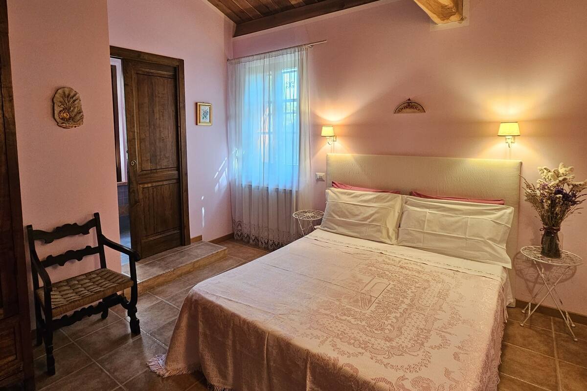 A bedroom with a wooden ceiling, a bed with a patterned bedspread, a bench, a door, and a window with curtains.