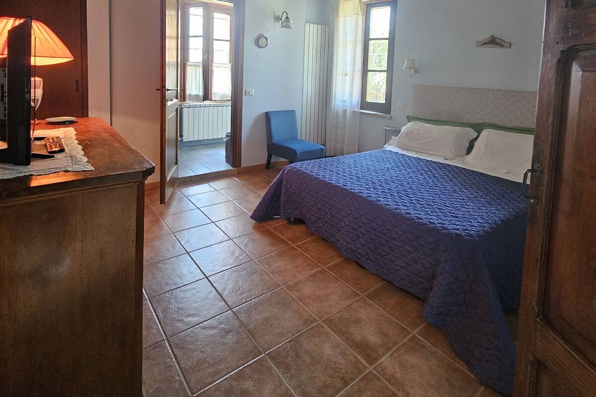 A bedroom with a wooden ceiling, a bed with a blue blanket, a desk with a lamp, and a chair.