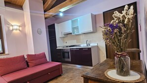 Una sala de estar acogedora con un sofá rojo, una mesa de comedor de madera y un florero con flores secas. Se ve una cocina con alacenas blancas y un salpicadero de azulejos al fondo.