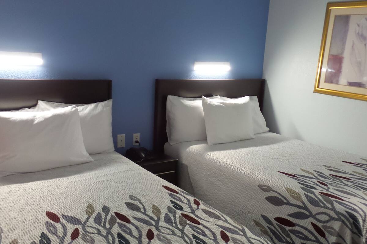 A hotel room with two beds, each with white bedding and a patterned duvet cover. There are bedside tables with lamps and a framed picture on the wall.