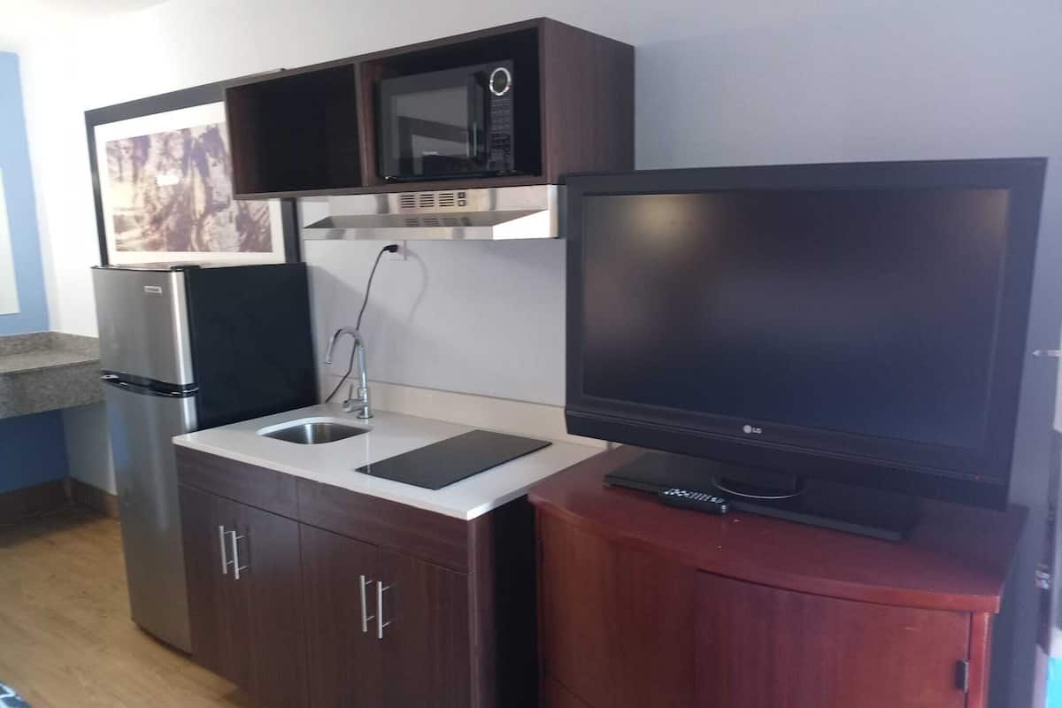 A compact kitchenette with a microwave, refrigerator, sink, and a flat-screen TV on a stand.
