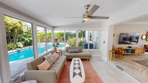 Un salon spacieux doté d’une grande fenêtre donnant sur une piscine, d’un ventilateur de plafond, d’un téléviseur et d’un coin repas avec des meubles modernes.