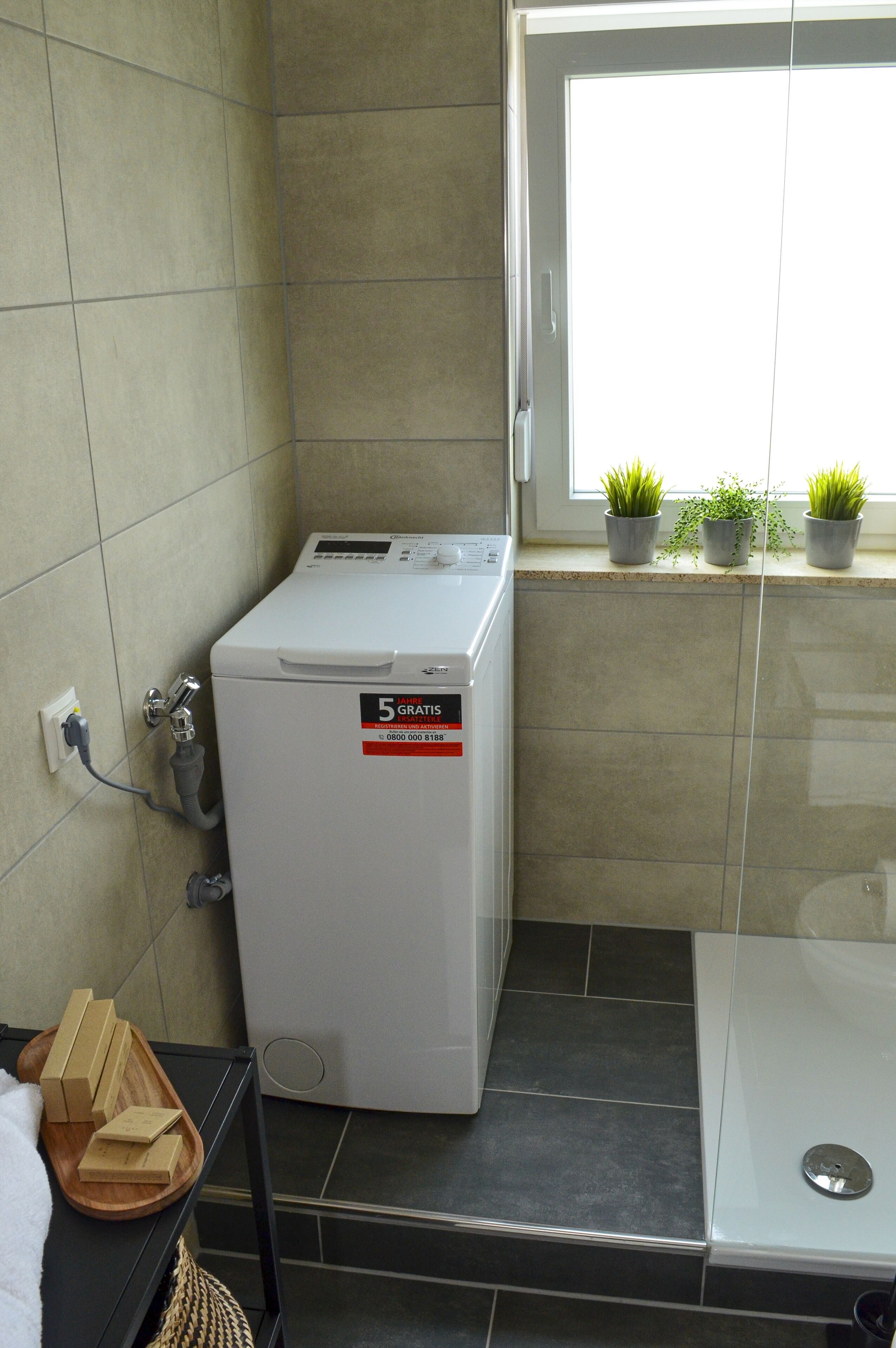 A compact laundry room with a front-loading washer and dryer, a small window with potted plants, and a tiled floor.