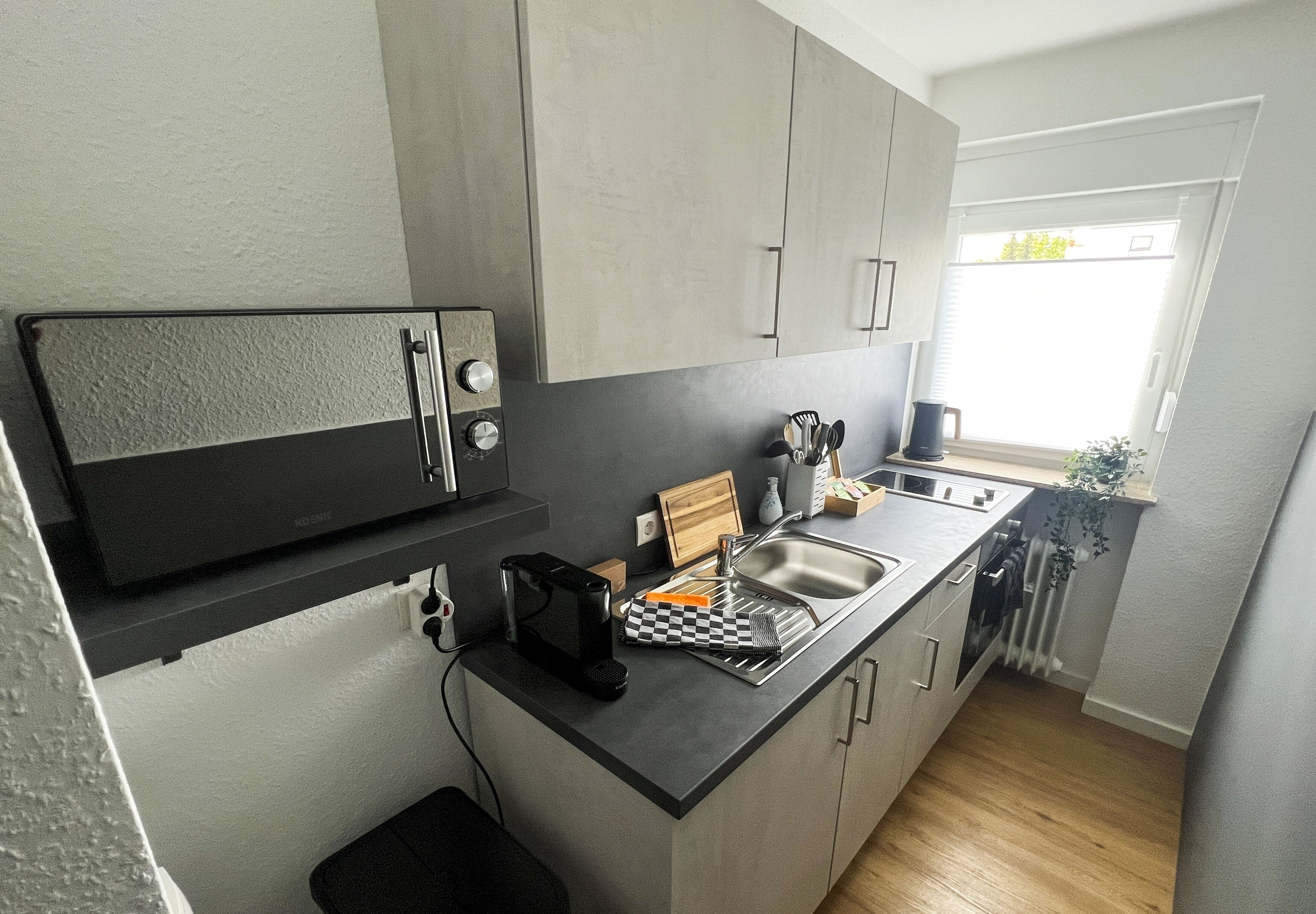 Cocina compacta con gabinetes grises, un microondas negro, un lavamanos y una ventana con vistas a la vegetación.