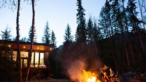 Eine Holzhütte, vor der ein Feuer brennt, umgeben von hohen Bäumen.