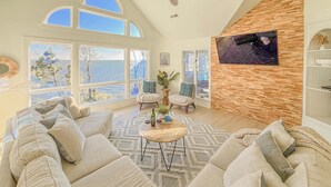 A spacious living room with a high ceiling, large windows, a ceiling fan, and a television mounted on a wooden wall. - Waterfront, Stunning Views + Pool + Kayaks! Pet friendly! (Corolla)