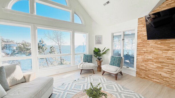 A spacious living room with large windows, a cozy sofa, two armchairs, a coffee table with a plant, and a wooden accent wall. - Waterfront, Stunning Views + Pool + Kayaks! Pet friendly! (Corolla)