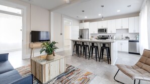 A modern living room with a TV, a coffee table with a plant, and a sofa. The kitchen has white cabinets, a bar area, and a dishwasher. - Charming Coastal Getaway: Explore and Relax in our 2 Bedroom Downtown Rental (Colonial Beach)