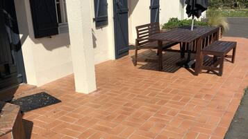 Un espace patio avec des carreaux de terre cuite, une table en bois et des bancs, ainsi qu’un parasol noir.