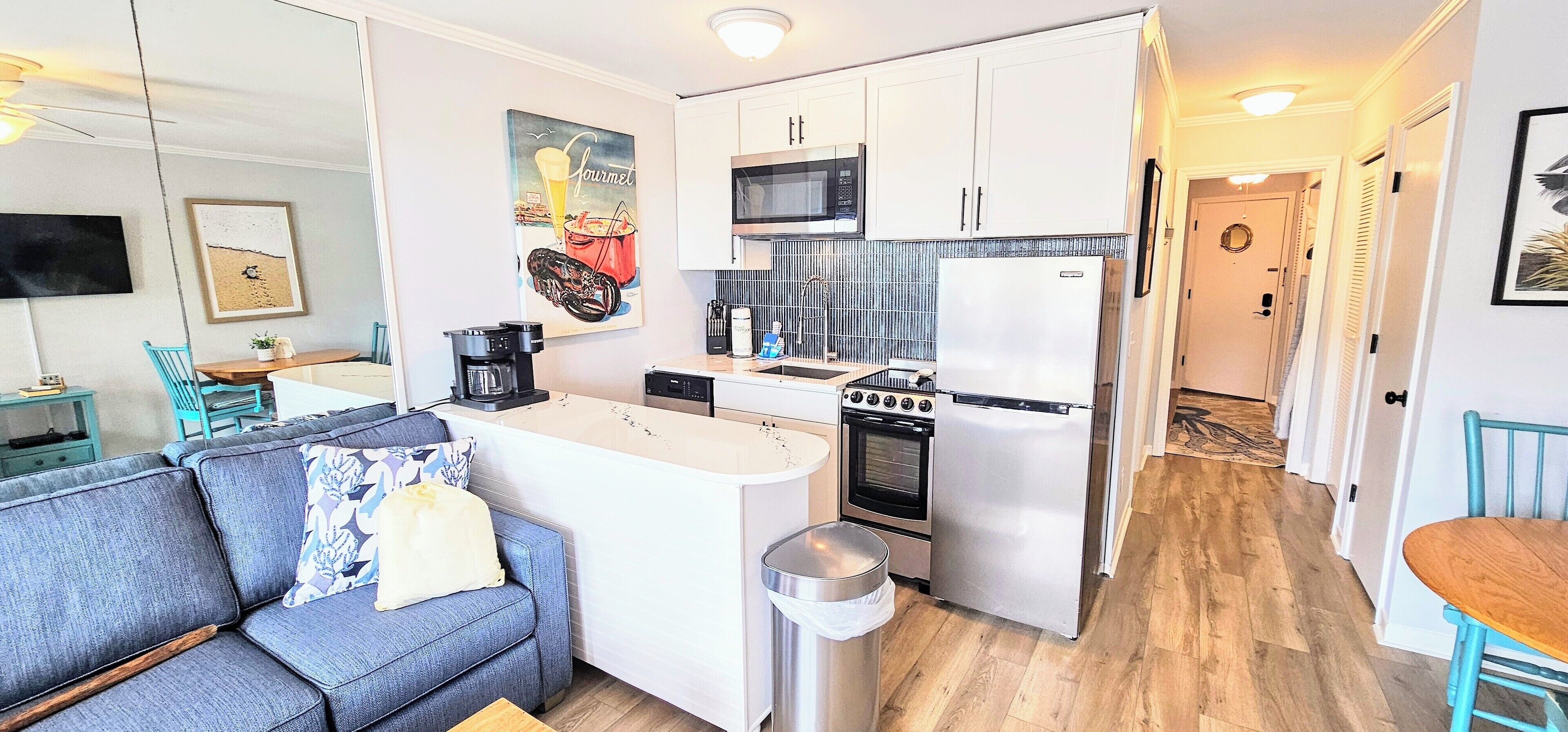 Una cocina compacta con armarios blancos, microondas, estufa y refrigerador. Hay un pequeño comedor con una mesa y sillas.