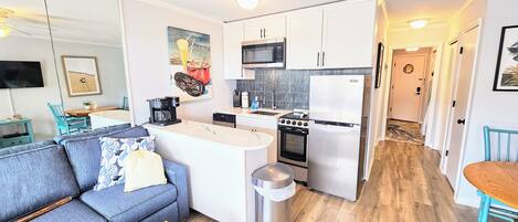 Una cocina compacta con armarios blancos, microondas, estufa y refrigerador. Hay un pequeño comedor con una mesa y sillas.
