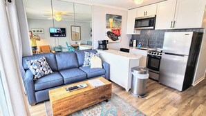 Un salon moderne avec un canapé bleu, une table basse en bois et un coin cuisine équipé d’appareils électroménagers en acier inoxydable.
