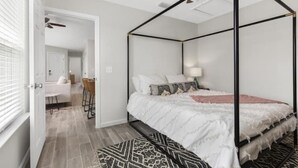A four-poster bed with a white bedspread and multiple pillows, a bedside table with a lamp, and a patterned rug on a wooden floor. - Stunning NEW / 2BR/Walk to Beach/Sleeps 5/Bunk (Panama City)